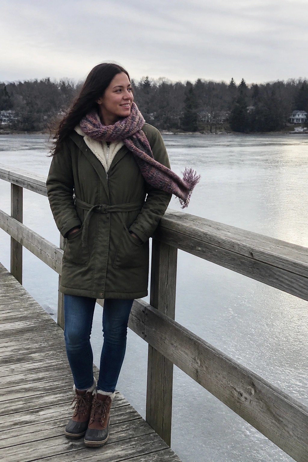 Woman in green parka and colorful scarf on wooden dock by frozen lake