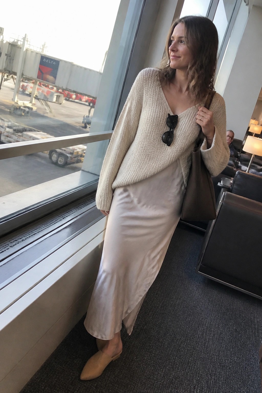 Woman in cream knit sweater and satin skirt holding bag at airport window.