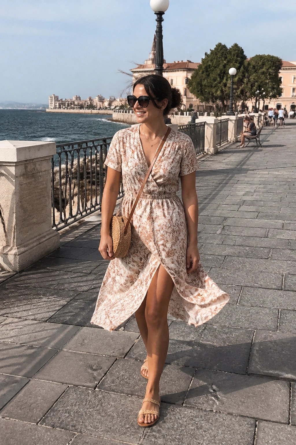 Woman in floral wrap dress walking seaside promenade with woven bag