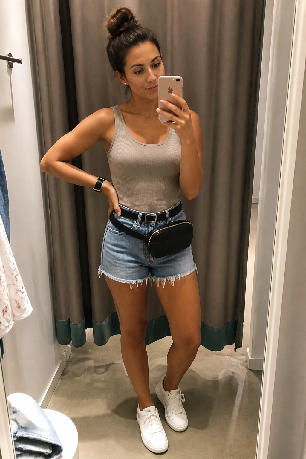 Woman in gray tank top and denim shorts taking mirror selfie in fitting room