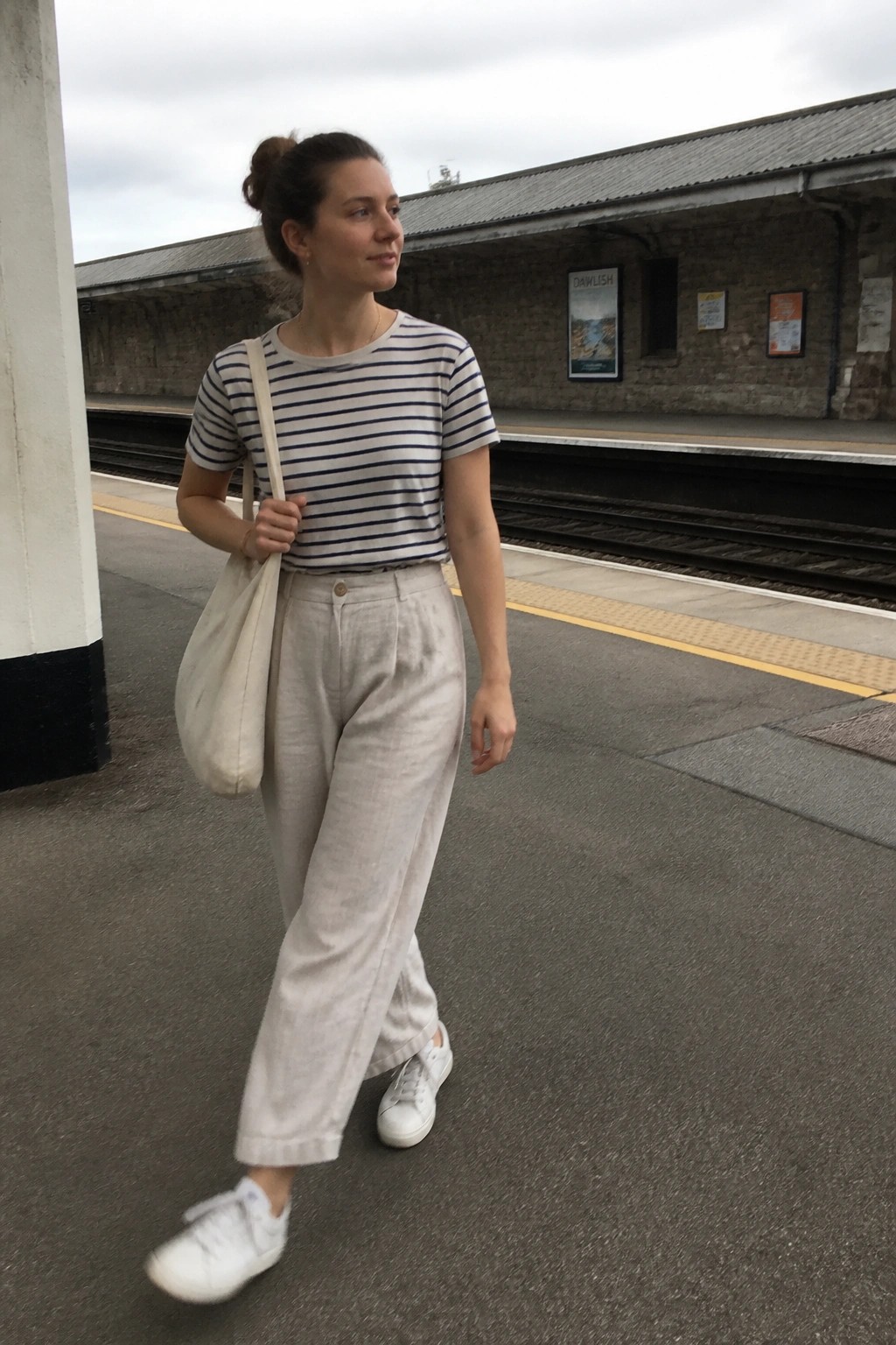 Woman in a navy striped t-shirt tucked into wide beige linen pants with white sneakers and a matching tote bag standing on a train platform