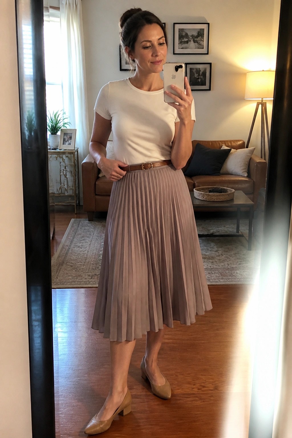 Woman in white top and pleated taupe skirt taking mirror selfie in living room