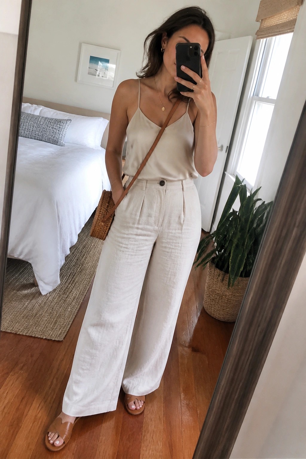 Woman in a cream camisole and white wide-leg linen pants with brown crossbody bag and slide sandals taking a mirror selfie indoors.