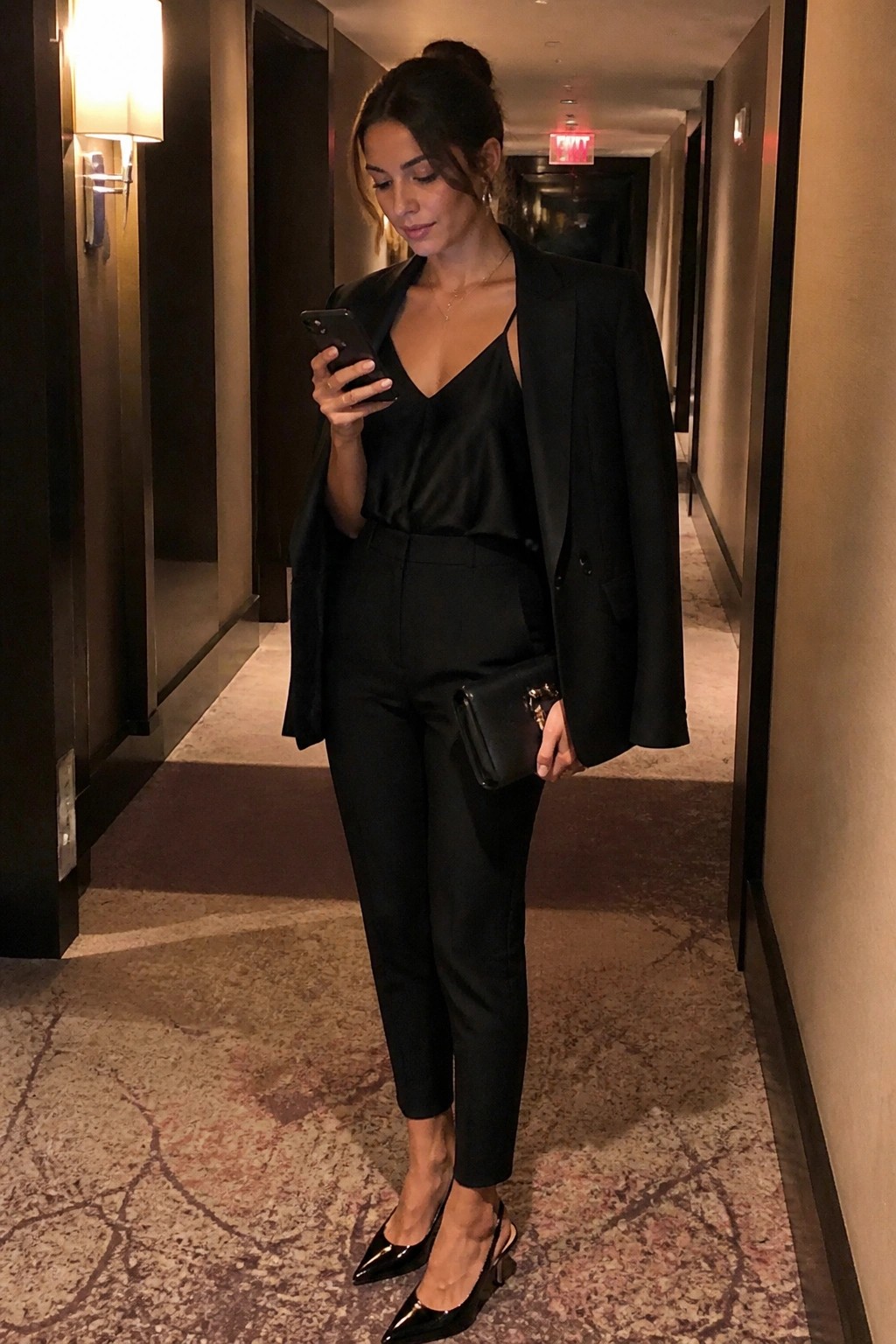 Woman in black blazer and satin top checks phone in hotel hallway