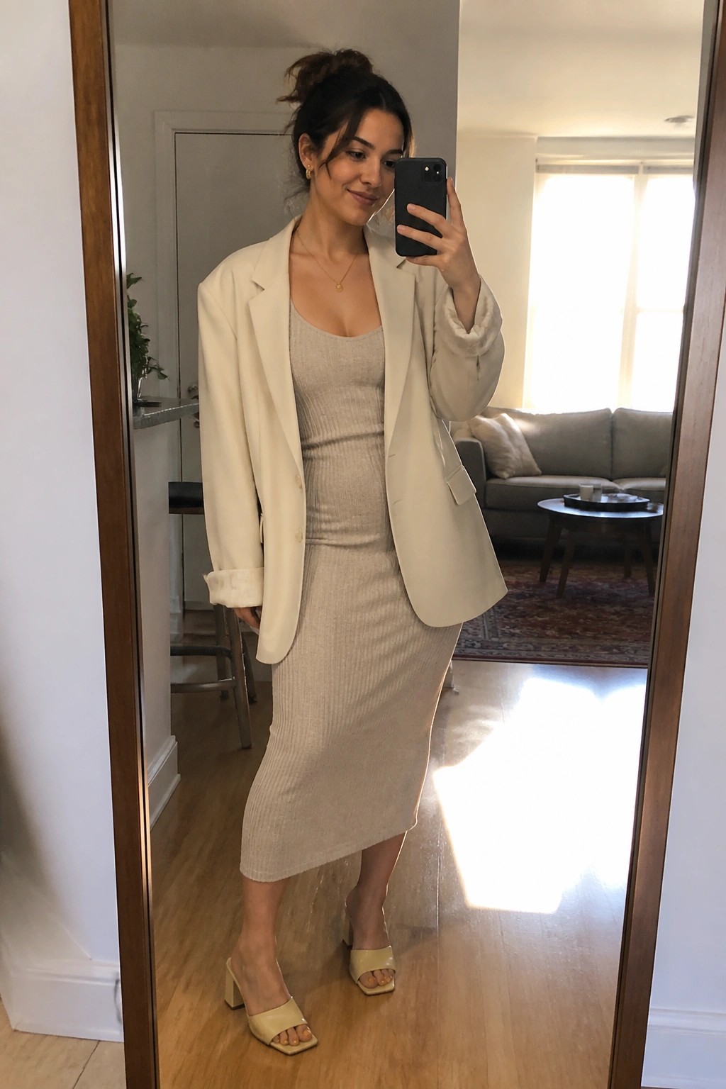 Woman in cream blazer and beige knit dress taking mirror selfie in living room