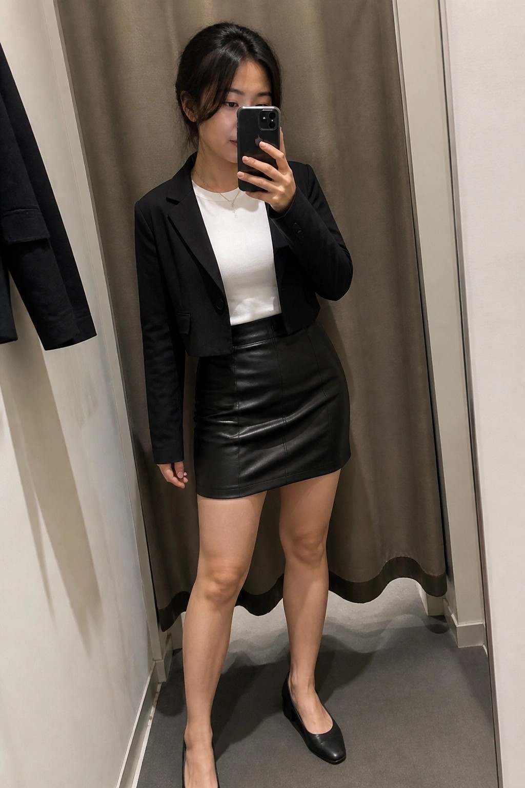 Woman wearing a black cropped blazer over a white top paired with a black leather mini skirt and black pointed flats while holding a phone for a mirror selfie.