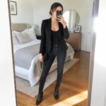 Woman in black blazer and jeans taking mirror selfie in bedroom.