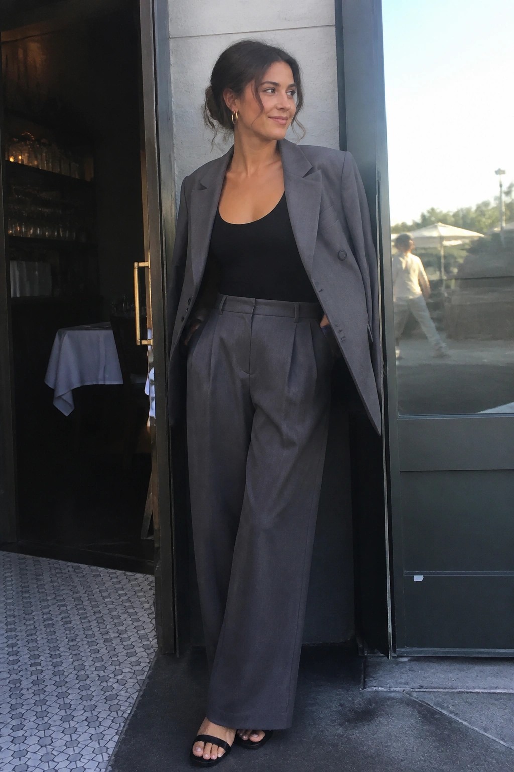 Woman in gray blazer and trousers posing in open cafe doorway, hands in pockets