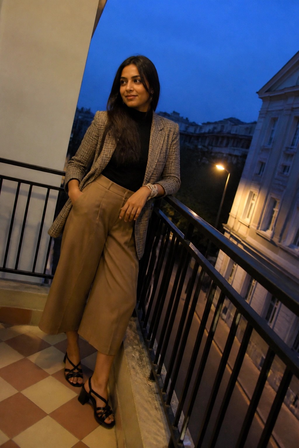 Woman in plaid blazer and wide-leg pants leaning on dusk balcony railing.
