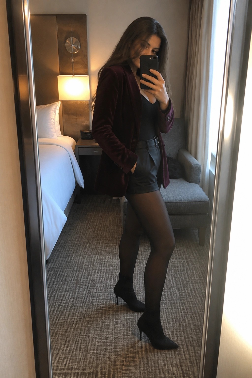 Woman in maroon velvet blazer over black bodysuit and leather shorts with black tights and pointed ankle boots posing for mirror selfie.
