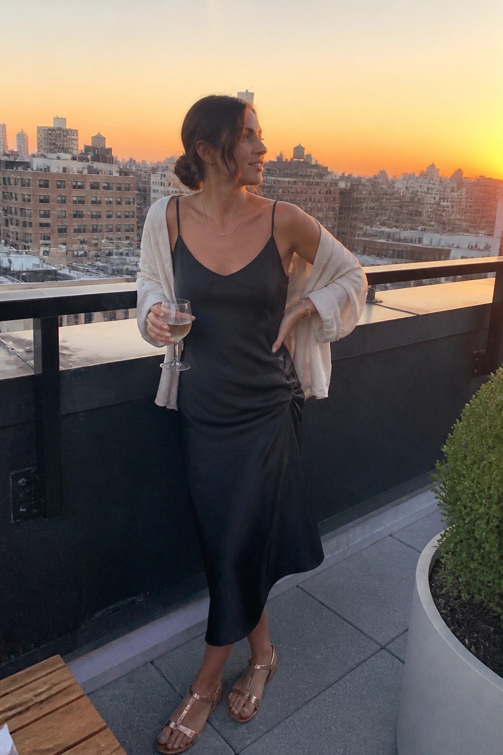 Woman in black satin midi slip dress with side ruching, cream cardigan draped over shoulders, thin gold necklaces, rose gold strappy sandals, standing on rooftop holding wine glass
