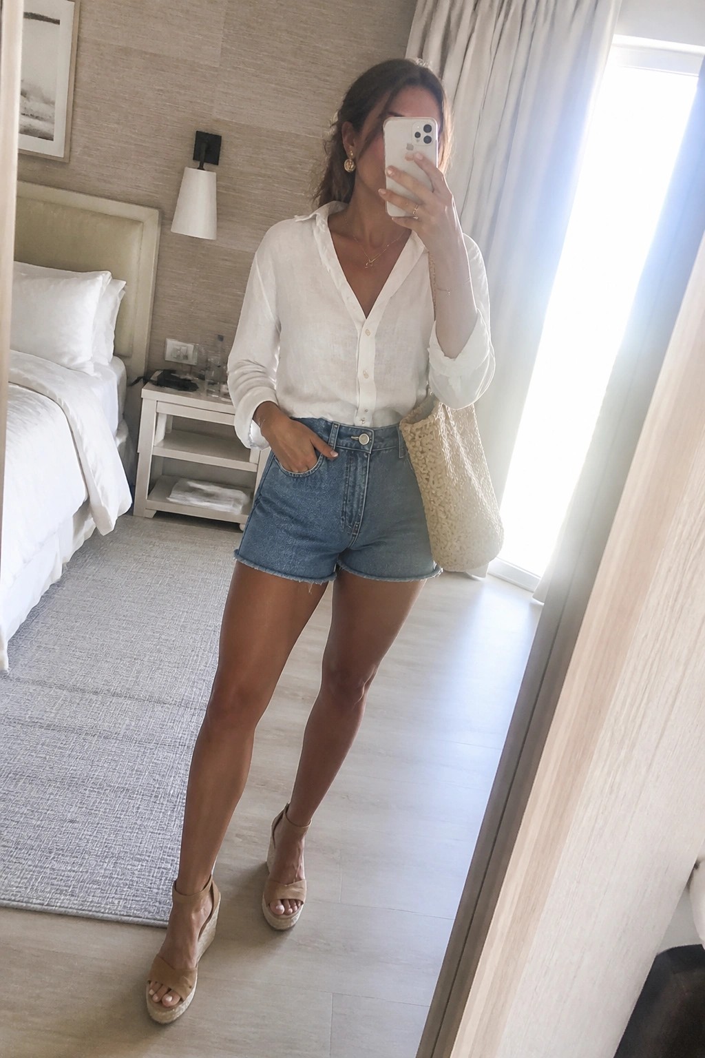 Woman in white shirt and denim shorts taking mirror selfie in hotel room