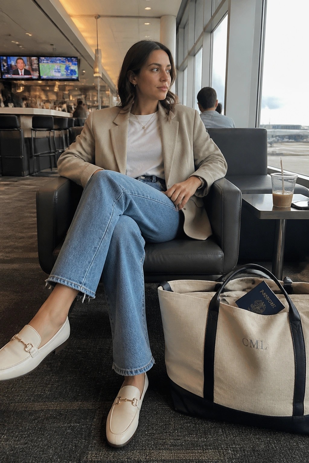 Woman in beige blazer and jeans seated in airport lounge with tote bag and passport