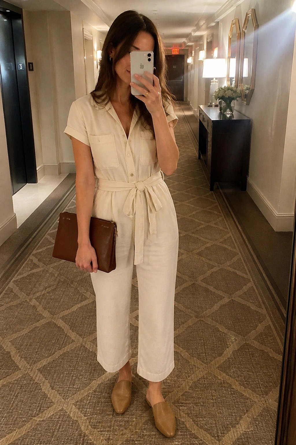 Woman in cream jumpsuit taking mirror selfie in hotel hallway with brown clutch.