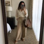 Woman in beige shirt and wide pants holding brown leather tote in mirror selfie
