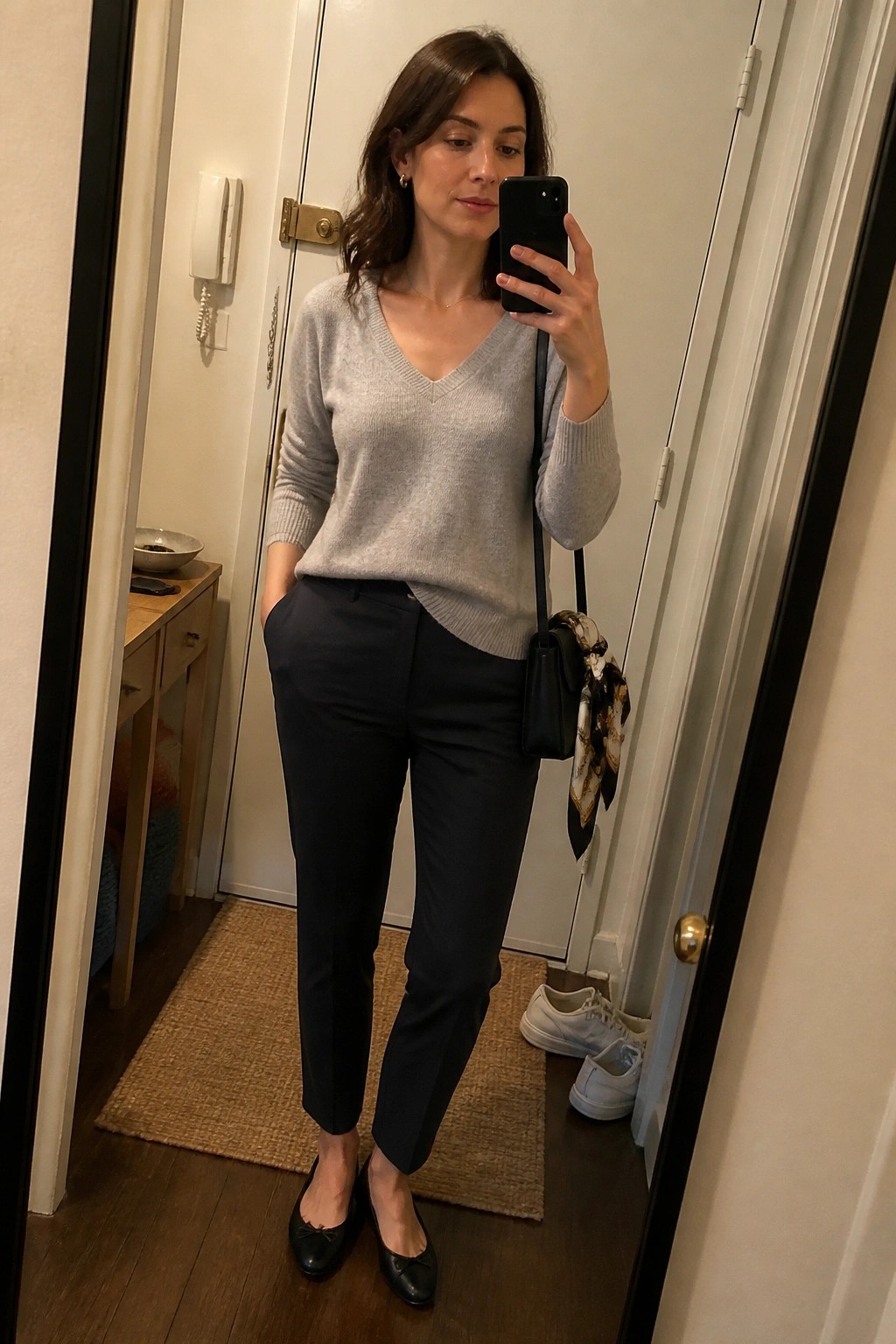 Woman in gray V-neck sweater and black pants taking mirror selfie indoors