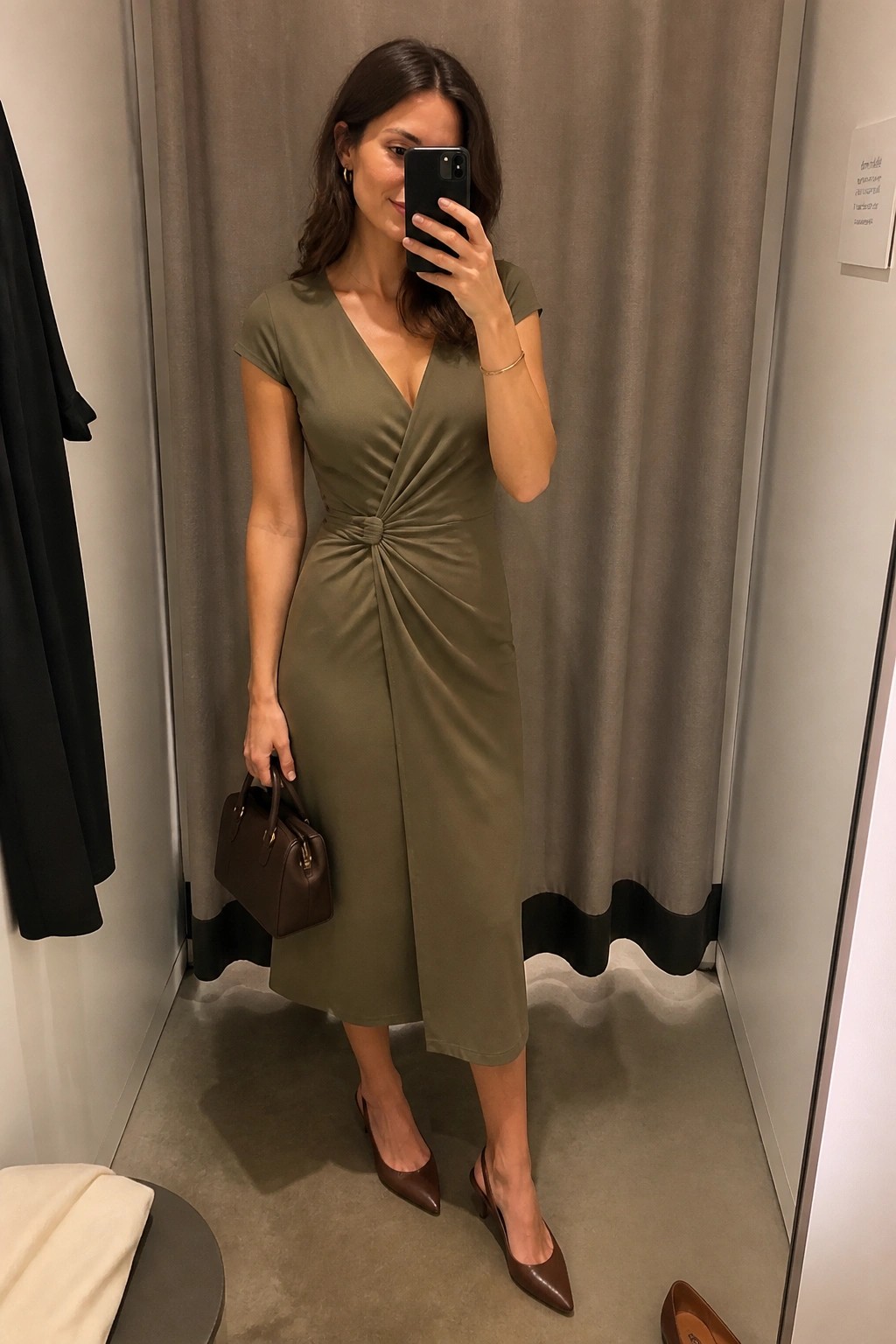 A woman taking a mirror selfie in a fitting room wearing an olive green midi dress with a knot detail at the waist, paired with brown slingback heels and a brown handbag.