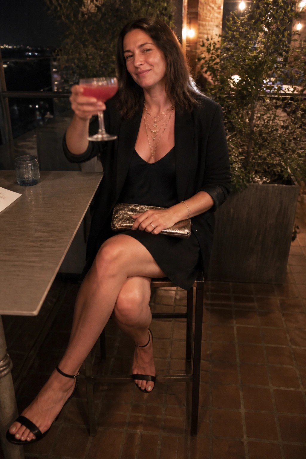 Woman seated wearing a black blazer over a black slip dress with layered gold necklaces a metallic clutch and black heeled sandals holding a pink cocktail.