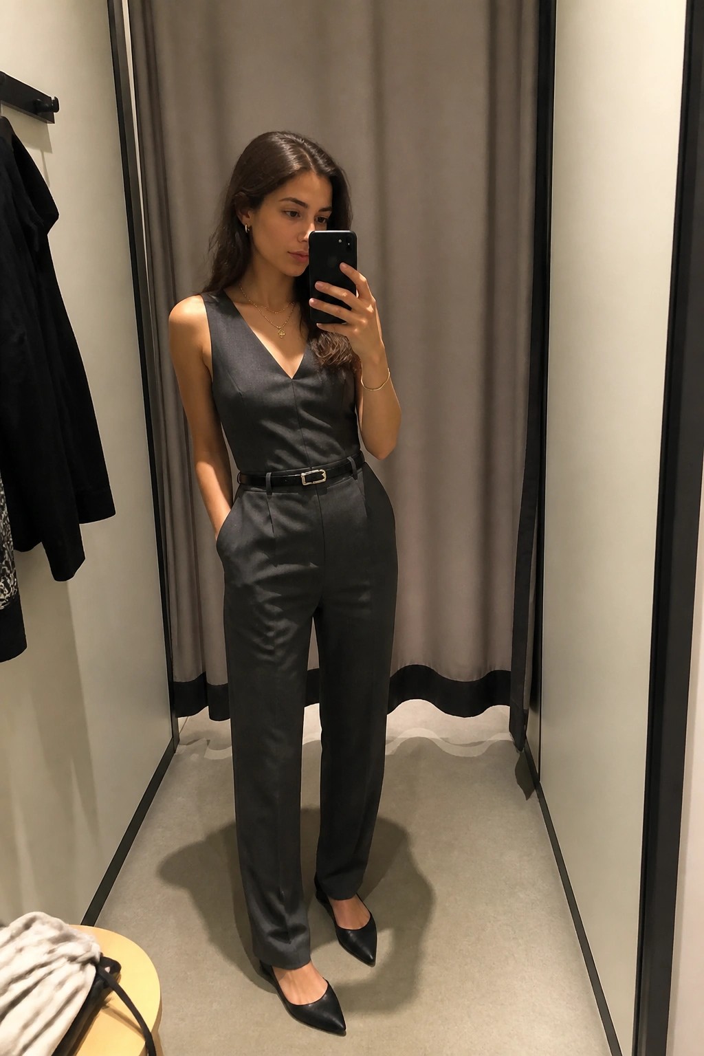 Woman in dark gray sleeveless jumpsuit taking mirror selfie in fitting room