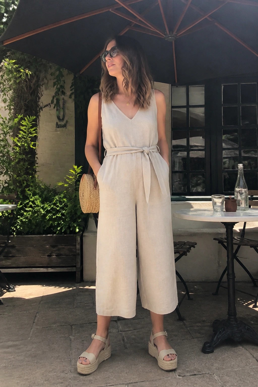 Woman in beige linen jumpsuit with woven bag under black umbrella at cafe patio