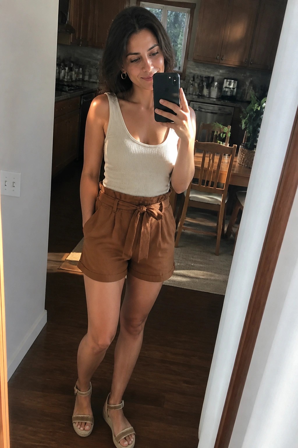 Woman in beige tank top and brown shorts taking mirror selfie indoors
