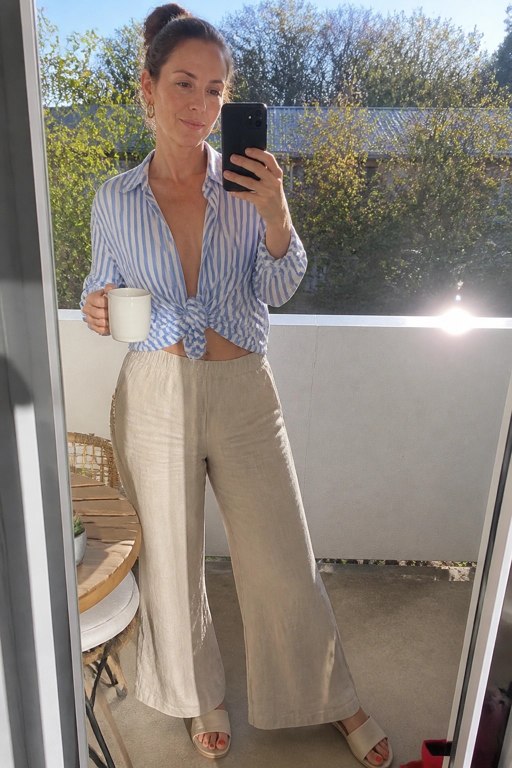 Woman in blue striped tied shirt and beige wide pants on sunny balcony with mug