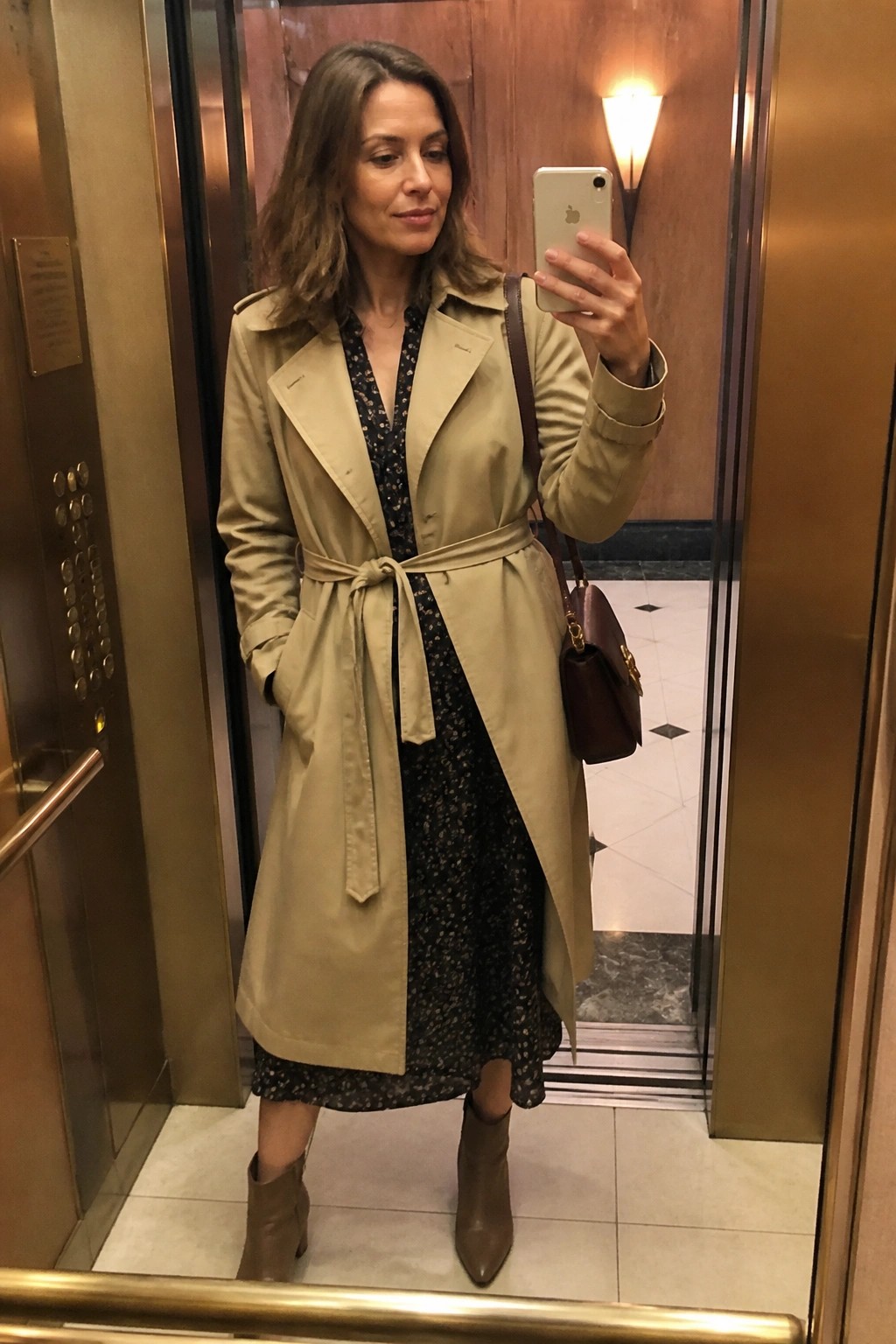 Woman wearing a belted beige trench coat over a black floral midi dress paired with taupe ankle boots and a brown leather handbag while taking a mirror selfie in an elevator.