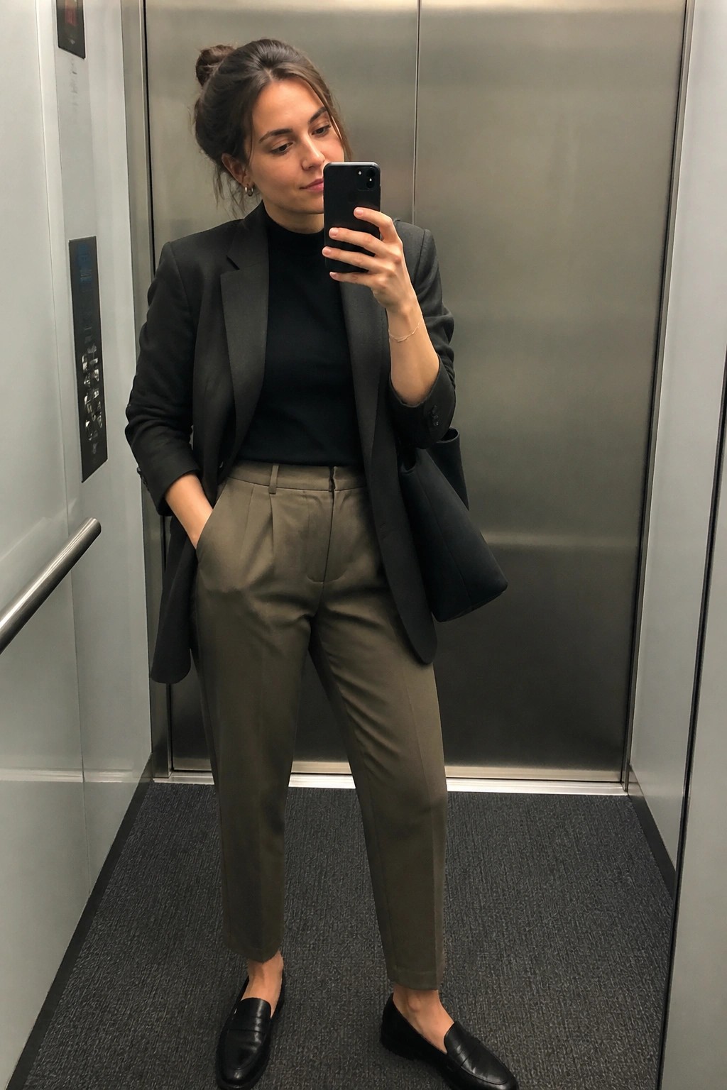Woman in black blazer over black turtleneck paired with olive green trousers and black loafers holding a black tote while taking a mirror selfie in an elevator.