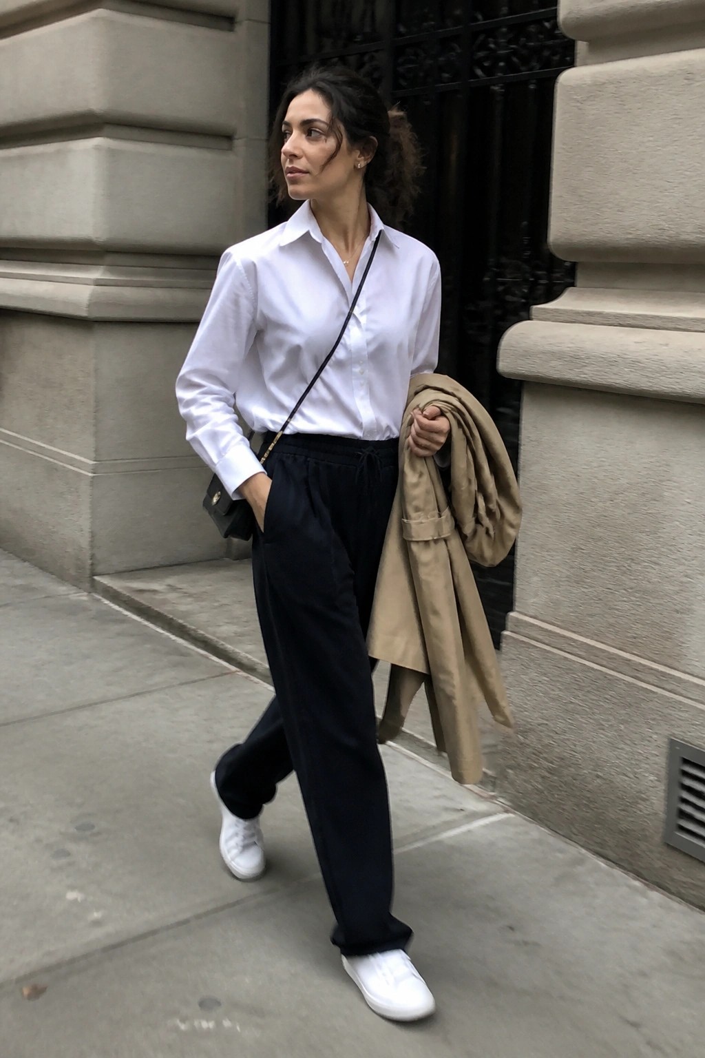 Woman in white button down shirt tucked into black drawstring pants paired with white sneakers while holding a beige coat and wearing a small black crossbody bag.