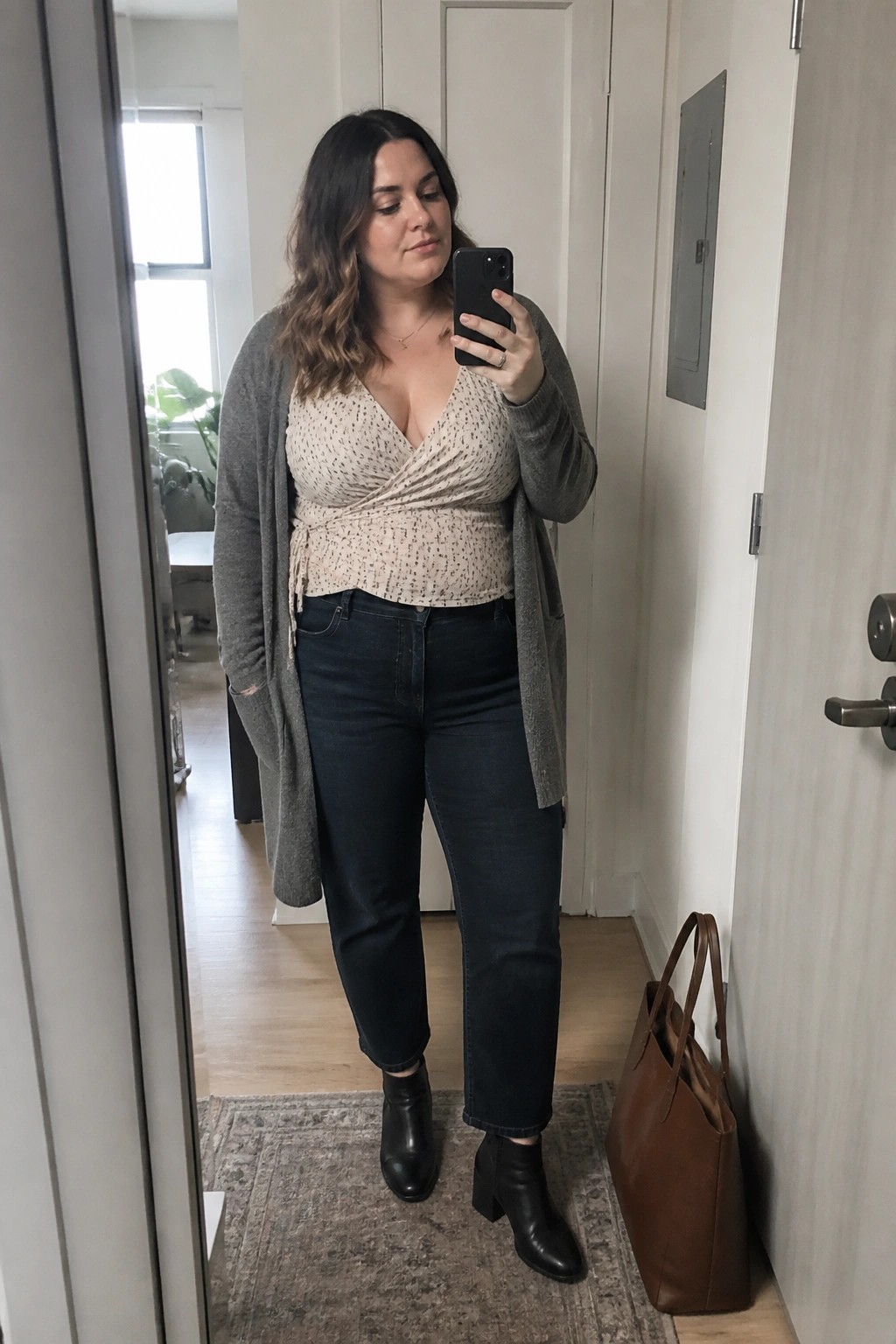 Woman in gray open cardigan over speckled wrap top paired with dark jeans and black block heel boots taking a mirror selfie.