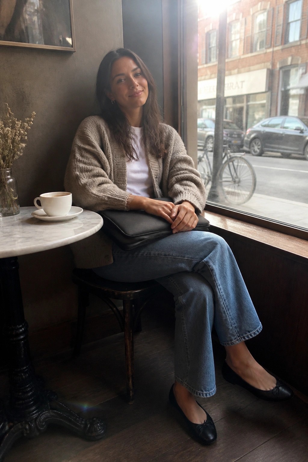 Woman seated by a window wearing a beige chunky knit cardigan over a white t-shirt, paired with blue wide-leg cropped jeans, black ballet flats, and holding a black leather clutch.