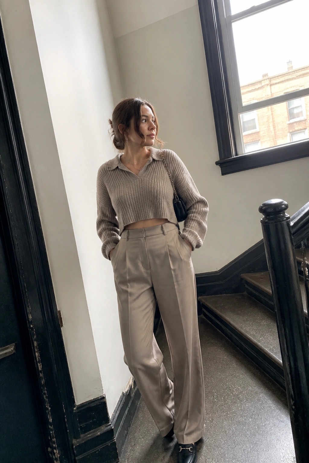 Woman wearing a beige cropped ribbed collared sweater tucked slightly into high waisted wide leg taupe trousers, paired with black loafers and a small black chain strap bag, standing indoors near stairs.