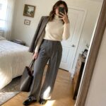 Woman in cream turtleneck and gray trousers with black blazer and tote bag