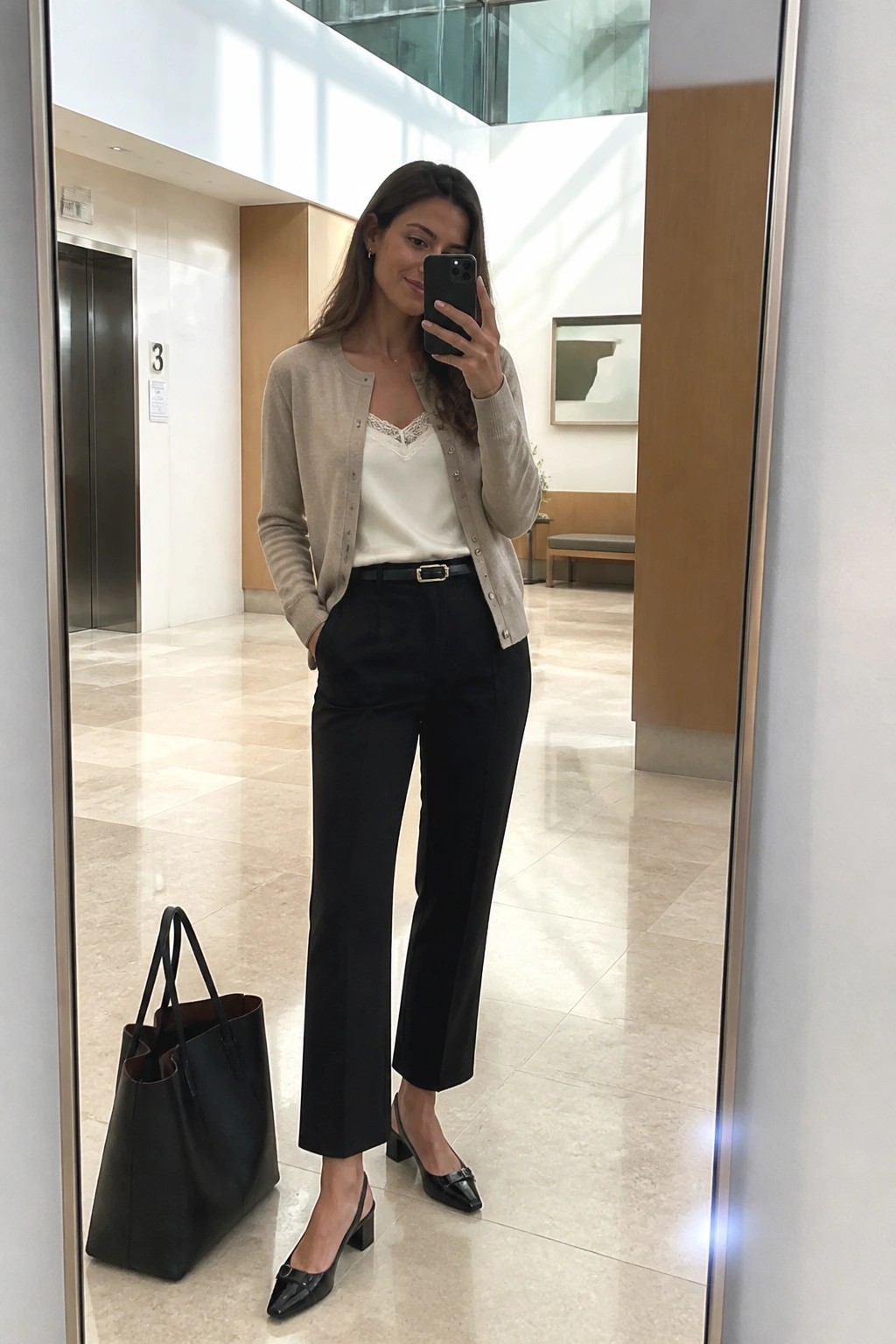 Woman wearing a beige knit cardigan over a white lace trim camisole with black straight leg trousers, black belt, and black slingback block heel shoes while holding a phone in a modern office lobby.