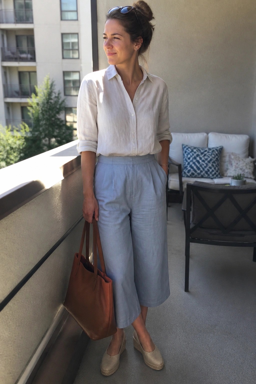 Woman in white shirt and light blue culottes holding brown tote on balcony