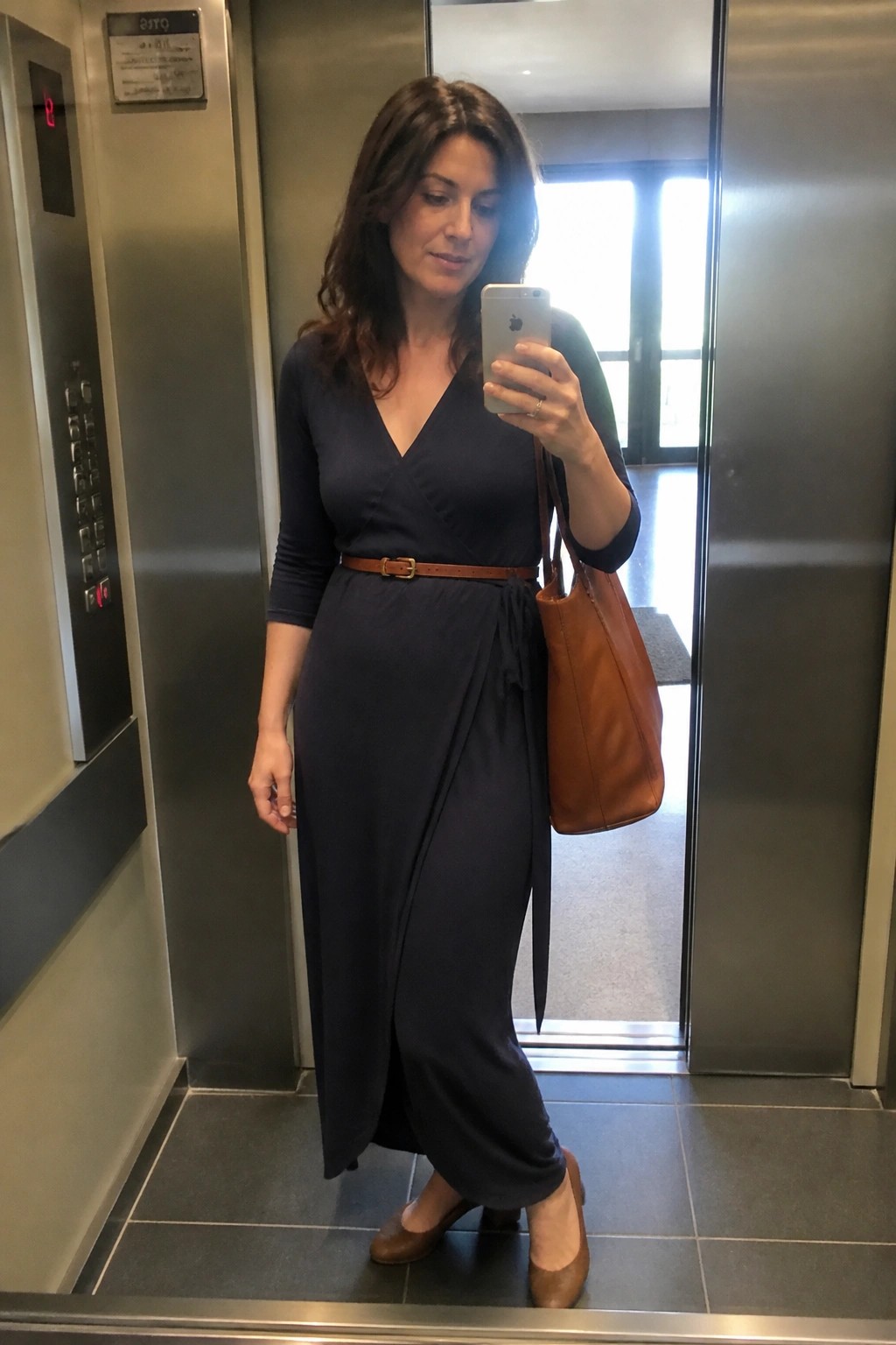 Woman in navy wrap dress taking mirror selfie in elevator with brown tote