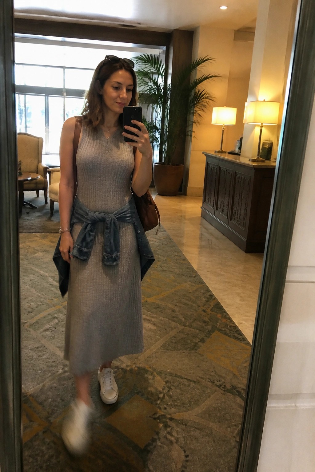Woman in gray knit dress with denim jacket tied at waist taking mirror selfie in lobby