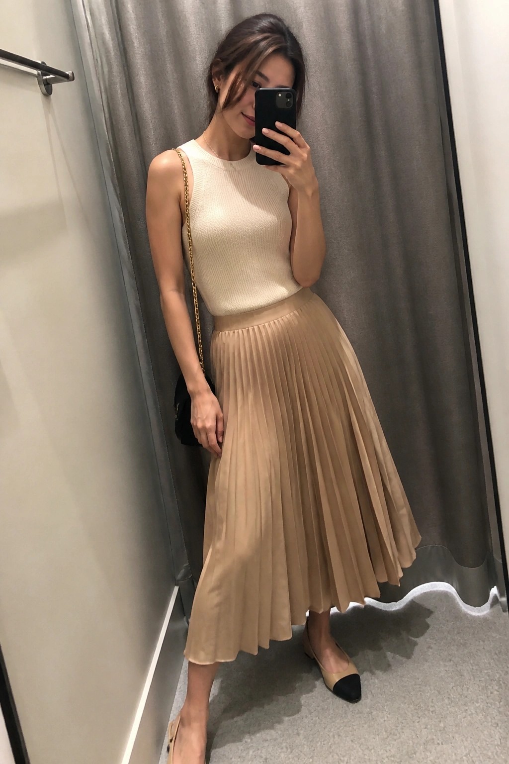 Woman in cream knit top and beige pleated skirt taking mirror selfie in fitting room