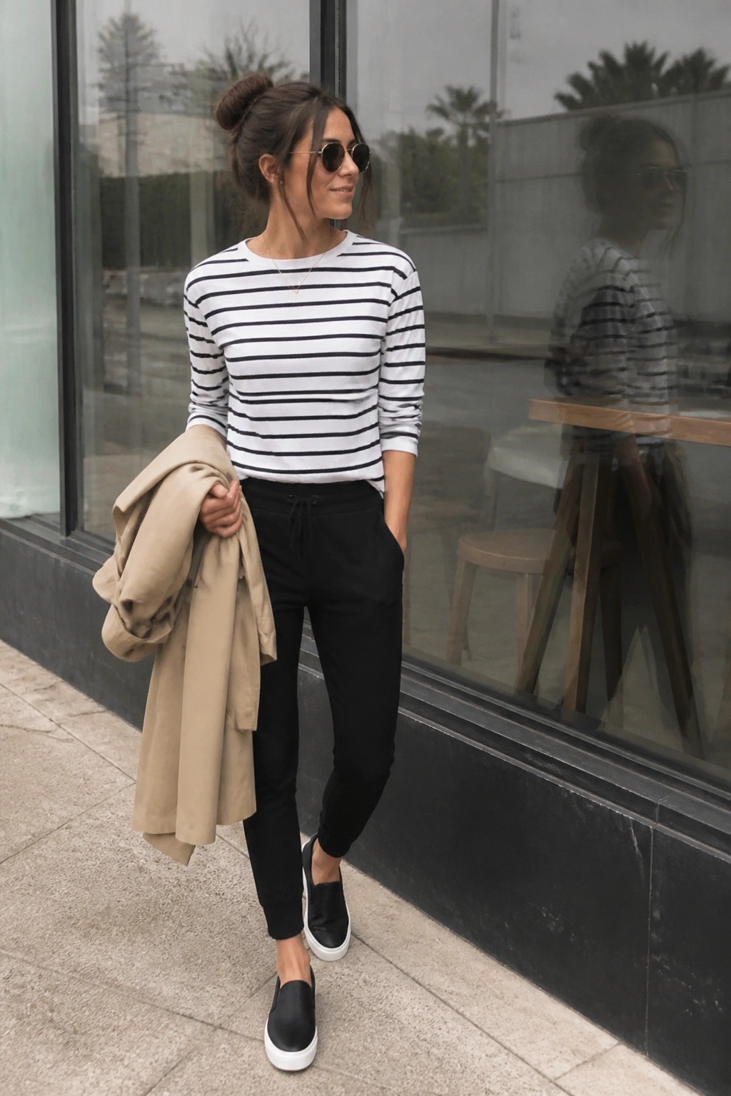 Woman wearing a black and white striped long sleeve top with black jogger pants, black slip on sneakers, and holding a beige jacket over one arm.