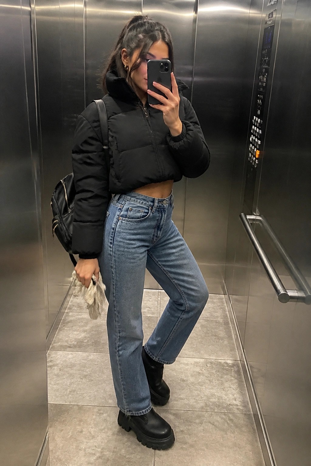 Woman in black cropped puffer jacket and jeans taking selfie in elevator