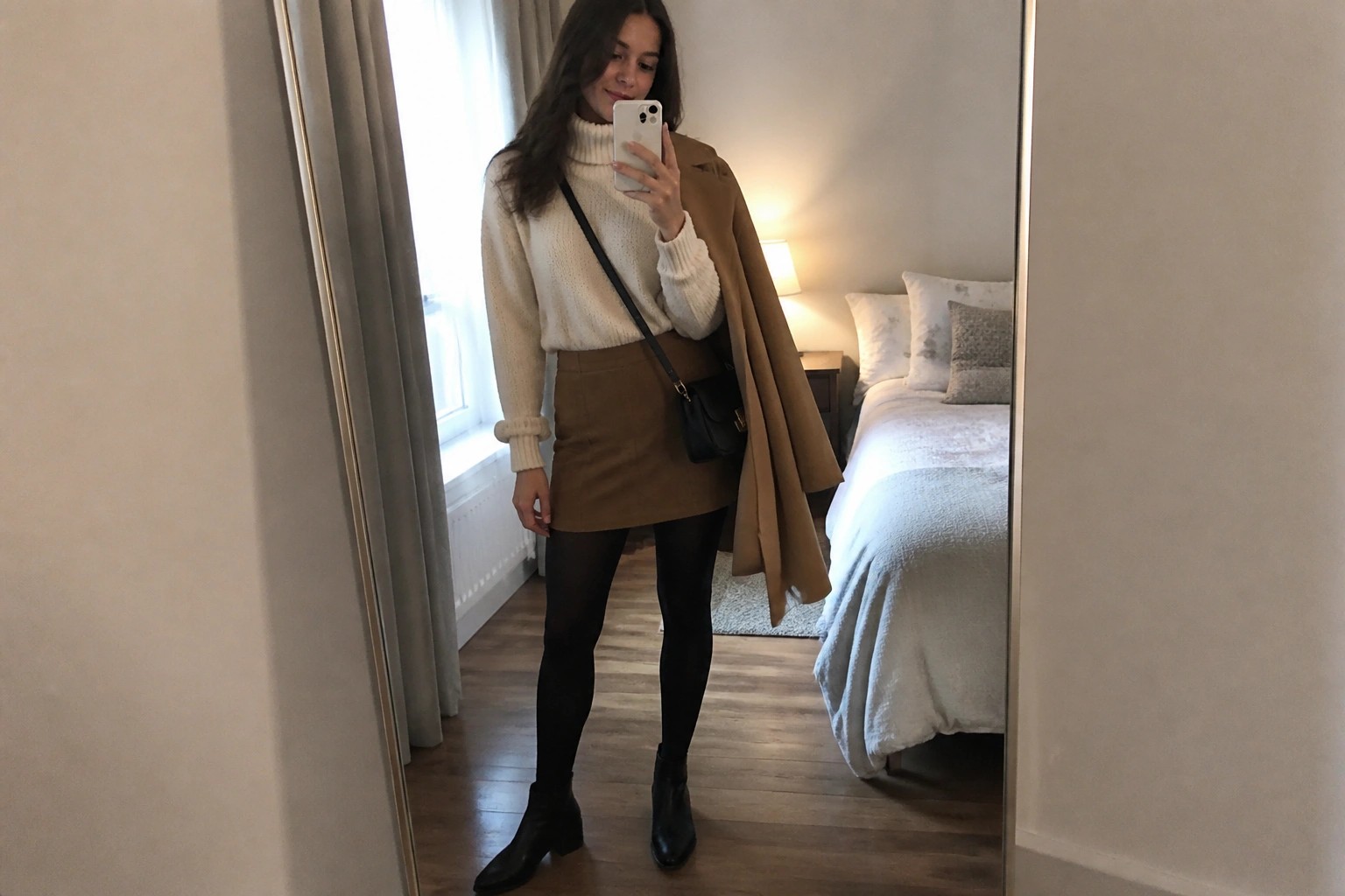 Woman in camel coat over cream sweater and brown skirt taking bedroom mirror selfie