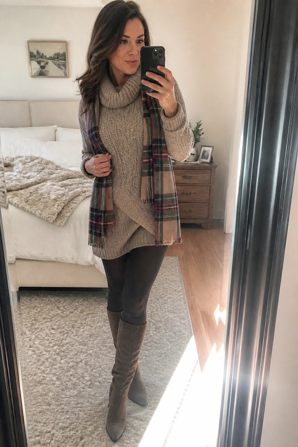 Woman wearing a beige cable knit turtleneck sweater draped with a plaid scarf in neutral tones black tights and taupe suede knee high boots while holding a phone for a mirror selfie.