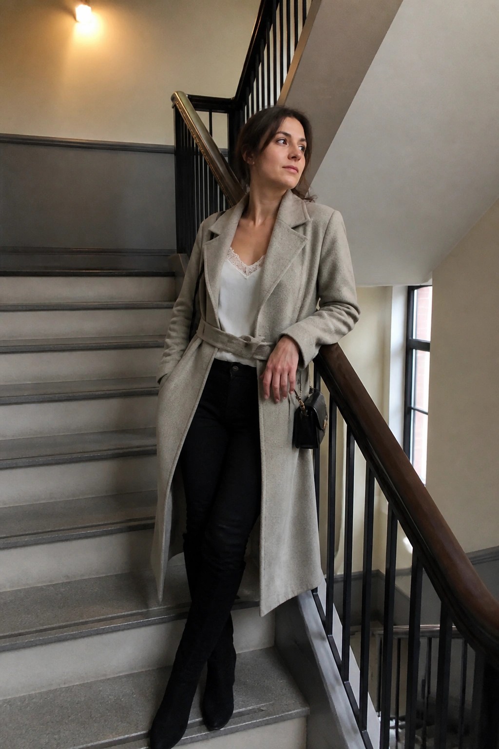 Woman in belted beige coat posing on indoor staircase with black handbag.
