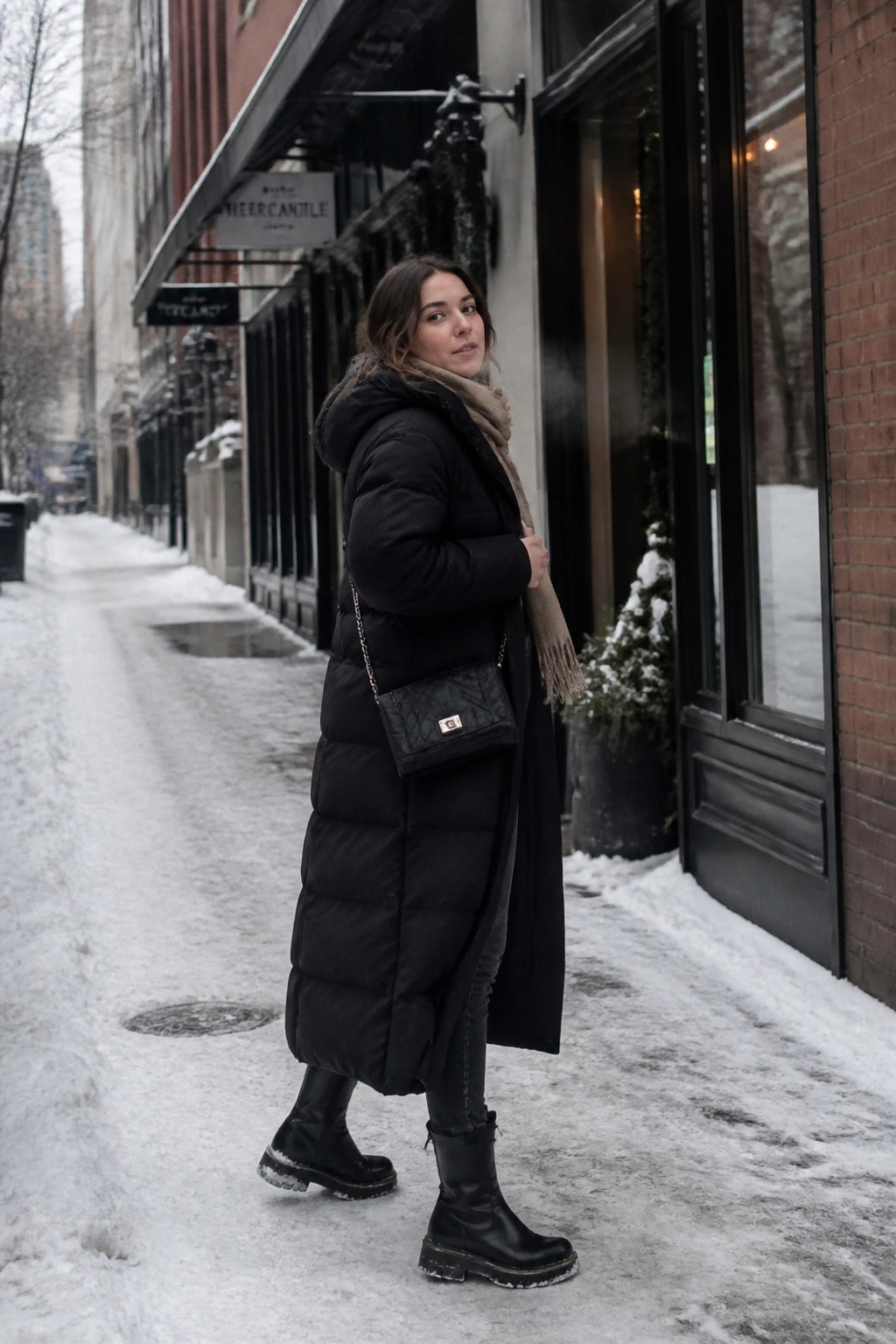 Woman in long black puffer coat and scarf on snowy sidewalk with handbag
