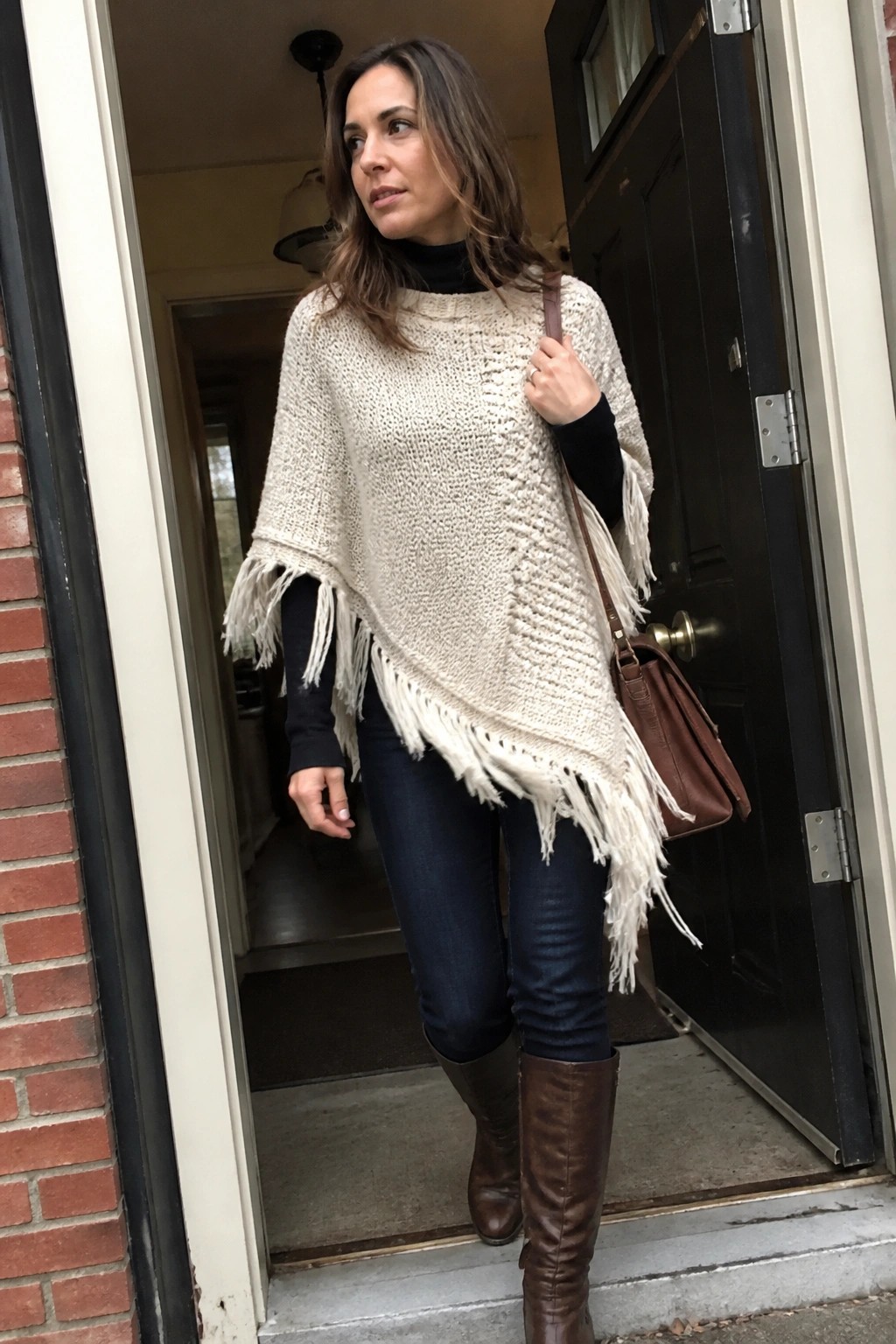 Woman in fringed cream poncho, blue jeans, and brown boots standing in doorway