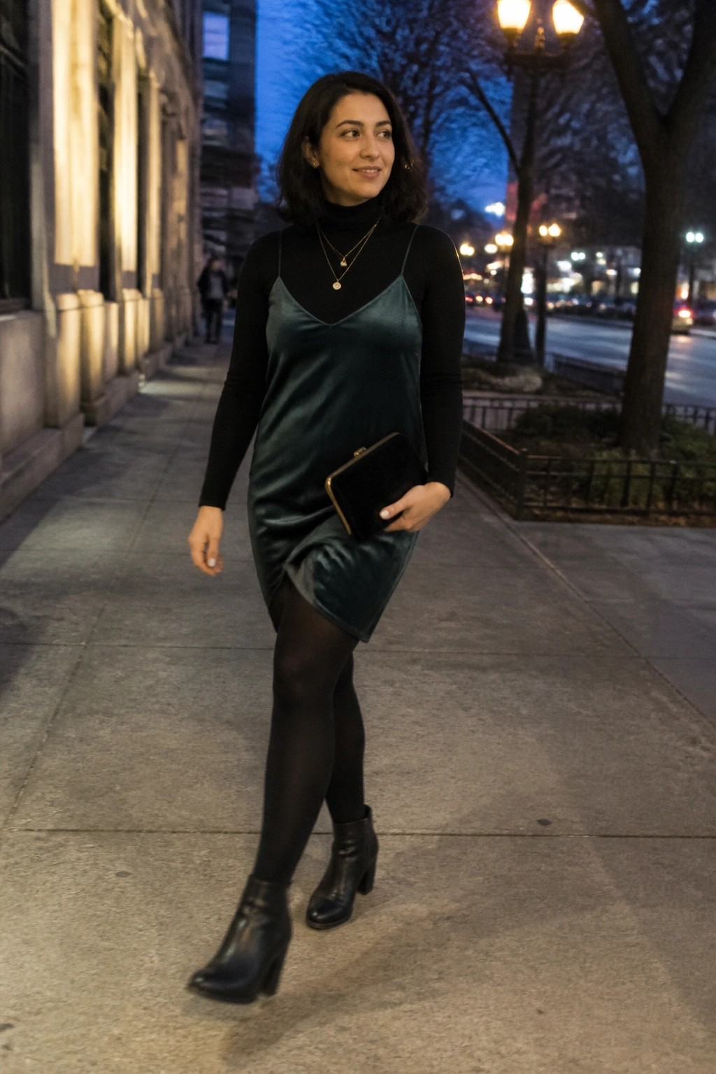A woman in a dark green velvet mini slip dress layered over a black turtleneck long sleeve top, black tights, black ankle boots, layered gold necklaces, and a black velvet clutch.
