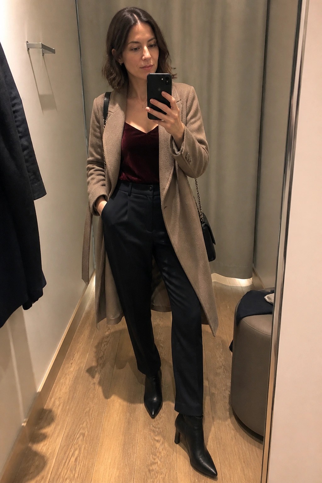 Woman in mirror selfie wearing long taupe coat over burgundy velvet camisole, black trousers, and black pointed ankle boots with chain strap handbag.