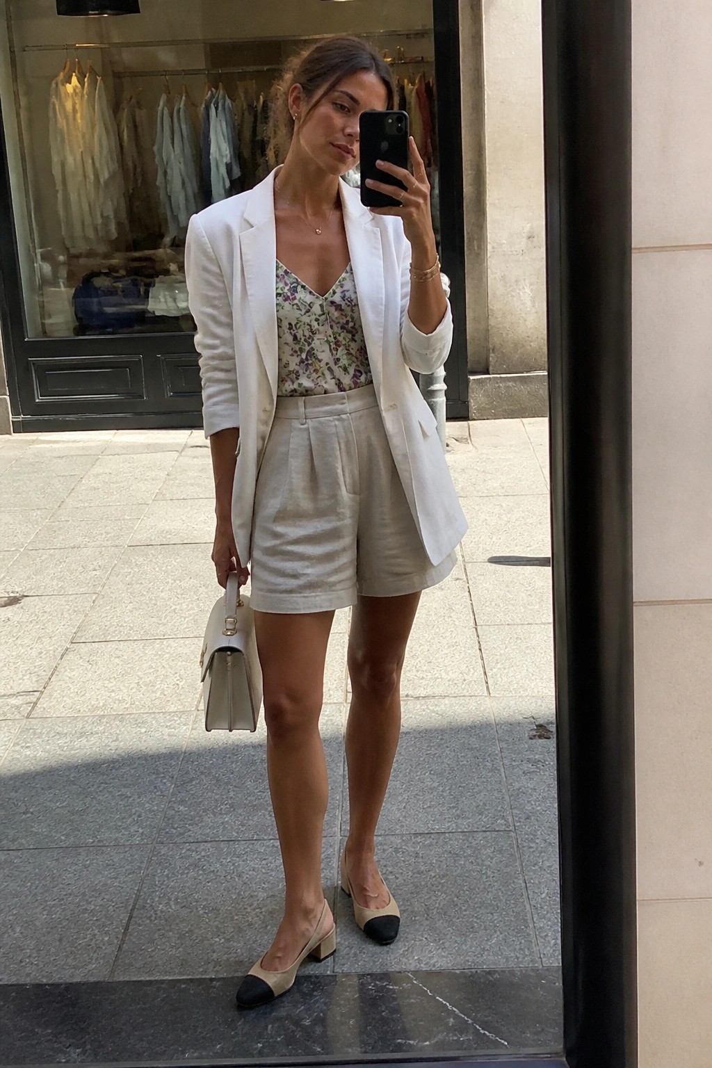Woman in white blazer and beige shorts taking mirror selfie with handbag.