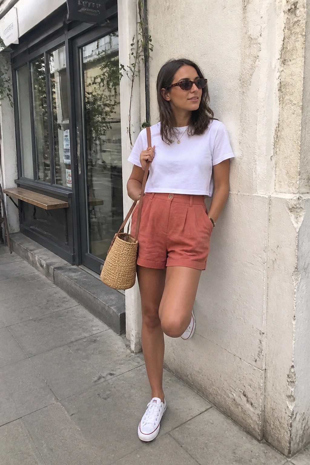 Woman in white crop top and salmon shorts holds woven bag against wall.