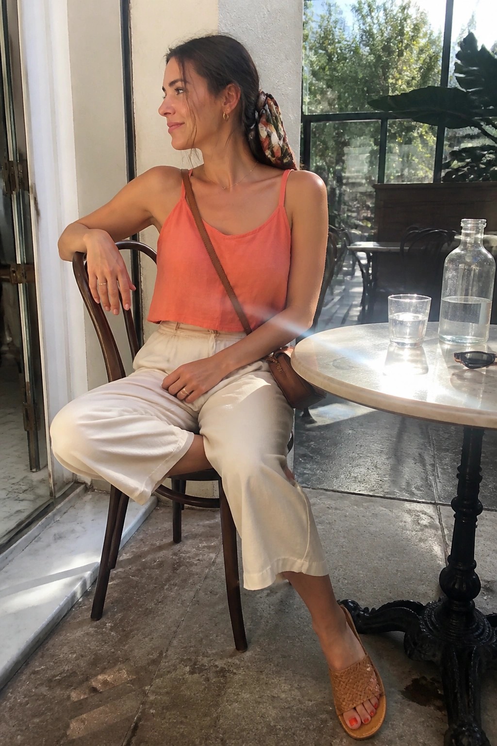 Woman in coral top and white pants seated at sunny outdoor marble cafe table